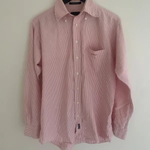 Rosa och vit randig skjorta Gant - Klassisk skjorta från Gant i rosa och vitt med tunna ränder. Skjortan har lång ärm, bröstficka och knappar framtill. Tillverkad i mjuk bomull och har en relaxed passform. Perfekt för dig som gillar en stilren och tidlös look.