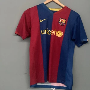 FC Barcelona Nike matchtröja - Klassisk FC Barcelona matchtröja från Nike med breda blå och vinröda ränder, broderat klubbmärke och Nike-logga i gult. Tröjan har korta ärmar, V-ringning och är tillverkad i lätt, ventilerande polyester. UNICEF-logga på bröstet. Perfekt för dig som älskar Barca!