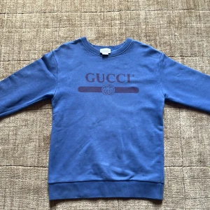 Blå tröja från Gucci - Snygg blå sweatshirt från Gucci med klassisk logga tryckt framtill i mörkare blått. Tröjan har rund halsringning och långa ärmar. Tillverkad i mjuk bomull som känns skön mot huden. Perfekt för dig som gillar stilrena designer och vill sticka ut med ett exklusivt märke. Använd 1 gång bara. En riktig bra deal om du frågar mig