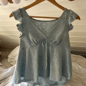Ljusblå blus med broderade hål och volang - Supersöt ljusblå blus med broderade hål och volangärmar. Toppen har v-ringning med knyt framtill och är ärmlös med romantiska detaljer. Perfekt för vår och sommar när du vill ha en luftig och feminin look.