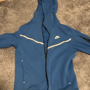Blå Nike hoodie med dragkedja - Blå hoodie från Nike med vit logga på bröstet och vita detaljer längs bröstet. Tröjan har huva, hel dragkedja och långa ärmar. Perfekt för en sportig look eller chill vardagsstil.