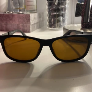 Svarta solglasögon Tommy Hilfiger - Snygga svarta solglasögon från Tommy Hilfiger med rektangulära bågar och mörkgula glas. Sidorna har en diskret röd detalj och logga. Lätta och stilrena, perfekta för att lyfta din look. Bra present till far, morfar/farfar eller till sig själv!