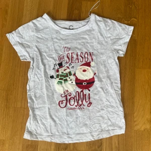 Julig t-shirt - Ljusgrå t-shirt från C i bomull med korta ärmar och tryck framtill av tomte och snögubbe samt texten 'Tis the season to be jolly'. Perfekt för dig som vill sprida julkänsla med en söt och rolig design.