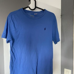 Blå t-shirt från Polo Ralph Lauren - Snygg blå t-shirt från Polo Ralph Lauren med klassisk broderad logga på bröstet. T-shirten är i mjuk bomull och har en enkel, stilren design med rund hals och korta ärmar. Perfekt för en clean och avslappnad look.