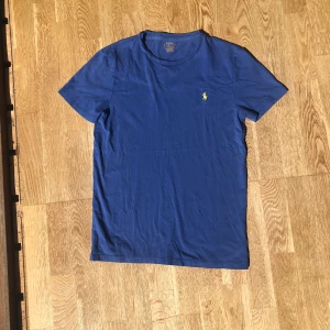 Blå t-shirt från Polo Ralph Lauren - Blå t-shirt från Polo Ralph Lauren i 100% bomull. Klassisk modell med korta ärmar, rund hals och gul broderad logga på bröstet. Slim fit för en snygg passform. Perfekt för dig som gillar stilrena och tidlösa plagg. Det är bara att skicka iväg ett meddelande om du vill veta någonting.
