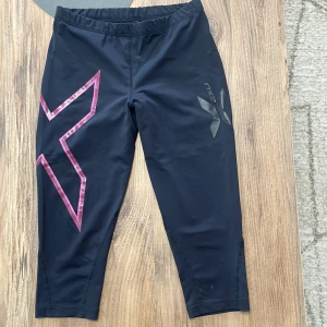 Svarta träningsbyxor från 2XU - Svarta träningsbyxor från 2XU med stor rosa X-detalj på ena benet och mindre svart X-logga på andra benet. Byxorna är trekvartsmodell med elastisk midja och tillverkade i ett stretchigt syntetmaterial som passar perfekt för träning.