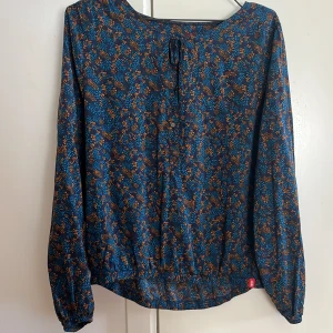 Blommig blus från Esprit i blått och orange - Säljer en blommig blus från Esprit i marinblått med orange och turkosa detaljer. Blusen har långa ärmar, knytning vid halsen och elastisk nederkant. Perfekt för dig som gillar färg och mönster! Storlek M.🌸