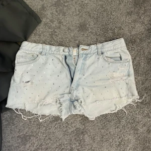 Ljusblå jeansshorts med nitar Gina Tricot - Snygga ljusblå jeansshorts från Gina Tricot med slitna detaljer, råa kanter och små silvriga nitar framtill. Klassisk femficksmodell och låg midja.
