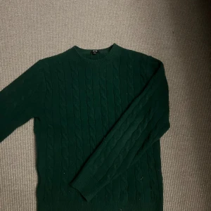 Mörkgrön stickad tröja från Uniqlo - Mörkgrön kabelstickad tröja från Uniqlo i storlek XS. Tröjan har rund halsringning, långa ärmar och ribbade muddar. Perfekt för dig som gillar klassisk stil med en twist. Materialet är mjukt och skönt, passar grymt till höst och vinter. Gjord av ull.