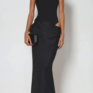 Svart långklänning med cut out-detalj - Supersnygg svart långklänning med figurnära passform och trendig cut out-detalj vid midjan. Klänningen har ärmlös design och hög halsringning, vilket ger en stilren och modern look. Perfekt för dig som vill sticka ut med en elegant men edgy stil.