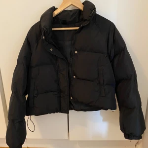 Svart pufferjacka från DAZY XS - Säljer en svart pufferjacka från DAZY i storlek XS. Jackan är kortare i modellen, har hög krage, dragkedja och tryckknappar framtill samt elastiska muddar vid ärmsluten. Perfekt för kalla dagar när du vill ha en stilren och enkel look.
