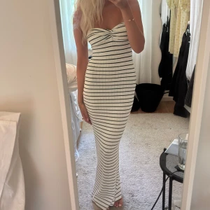 Vit långrandig bandeauklänning - Supersnygg lång bandeauklänning i vitt med svarta horisontella ränder. Klänningen är figurnära och har twistad detalj vid bysten. Materialet är ribbat och stretchigt, perfekt för en trendig look.