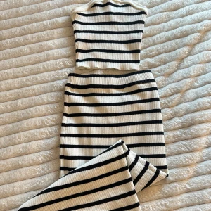 Randig ribbad långkjol i stickat set - Säljer en snygg ribbstickad långkjol med topp i vitt med svarta horisontella ränder. Kjolen är figurnära och har en liten slits i sidan. Materialet är mjukt och stretchigt stickat, perfekt för en trendig look. 