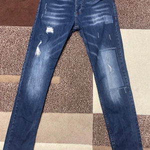 Dsquared2 blå jeans  - Snygga blå jeans från Dsquared2 med slim passform och slitna detaljer framtill. Jeansen har klassisk femficksdesign, knappar med Dsquared2-logga och tydliga slitningar och patchar för en trendig look. Perfekta för dig som gillar streetstyle. Dem är äkta och köptes inte så länge sen.