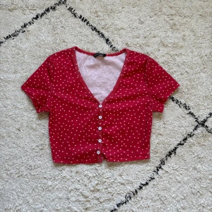 Röd croppad topp med hjärtan från SHEIN - Supersöt röd croppad topp från SHEIN med små vita hjärtan över hela. Toppen har korta ärmar, v-ringning och knappar framtill. Perfekt för dig som gillar gulliga detaljer och vill ha en topp som sticker ut lite extra.