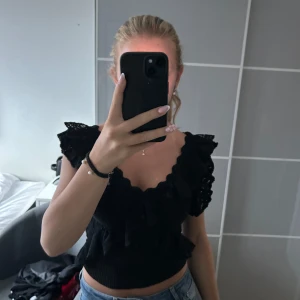 Svart blus med volang och spets - Supersöt svart blus med volangdetaljer och spets på axlarna. Djup v-ringning med vågig kant och croppad passform. Perfekt att styla med jeans eller kjol för en trendig look.