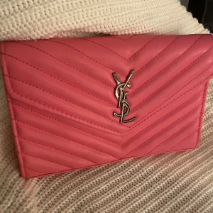 Rosa YSL kuvertväska i skinn - Säljer en snygg rosa kuvertväska från YSL i quiltat skinn med silverfärgad logga framtill. Väskan har ett stilrent kuvertformat lock och är perfekt att bära i handen. Supersnygg detalj till din outfit!