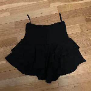 Svart volangkjol  - Supersöt svart kortkjol med volanger och smockad midja. Kjolen har en flowy känsla och passar perfekt till sneakers eller boots. Enkel att styla och ger en cool vibe till din outfit.