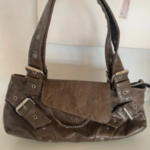 Brun axelväska med kedjedetalj - Cool brun axelväska i skinnimitation med snygga metalldetaljer, spännen och kedja framtill. Väskan har breda axelband med nitar och öglor, samt dragkedja på baksidan. Perfekt för dig som gillar en edgy stil och vill ha plats för allt du behöver.