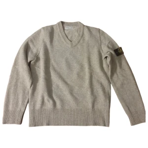 Beige/grön v-ringad Stone Island tröja - Snygg beige v-ringad tröja från Stone Island med klassisk patch på ärmen. Tröjan är stickad i mjuk ull och har ribbade muddar vid ärmslut och nederkant. Perfekt för dig som gillar stilren och clean streetwear. Köpt utomlands för ca 1 år sedan, knappt använd och inköpspriset var runt 10 tusen.