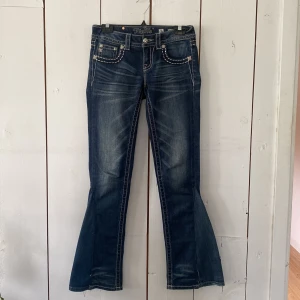 Miss Me blå bootcut jeans strl 26 - Snygga blå bootcut jeans från Miss Me med kontrastsömmar och stora broderade detaljer med nitar på bakfickorna. Jeansen har låg midja och klassisk femficksdesign. Det är även en insydd kil (bild 5) som den förra ägaren sytt som inte är jättebra sydd. Måttet på midjan hela vägen runt är 81cm. Skicket på jeansen är överlag bra bortsett från den insydda kilen.