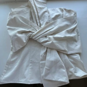 Vit axelbandslös topp med stor rosett - Superfin vit axelbandslös topp med en stor, iögonfallande rosett framtill. Snygg bandeau-modell som sitter tight och markerar midjan. Perfekt för dig som vill sticka ut med en elegant och trendig look.