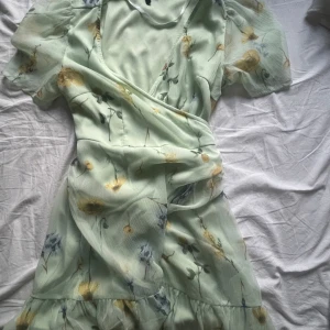 Blommig kort klänning  - Superfin mintgrön kortklänning från Vero Moda med blommigt mönster i gult och blått. Omlottmodell med knyt i midjan, volang nertill och korta puffärmar. Perfekt för sommarens alla tillfällen! 