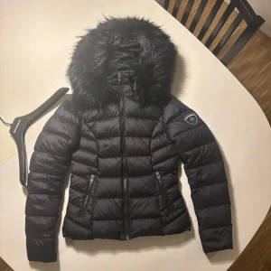 Svart dunjacka från Rockandblue med fuskpäls - Jättefin helt oanvänd svart dunjacka från Rockandblue i storlek 34 med fluffig svart fuskpäls som även går att ta av. Jackan har dragkedja framtill och två innefickor med dragkedja. Galjen till den finns även kvar! Säljer på grund av att den var för liten vid köp! Går att spänna in vid midjan baktill därav knapparna på baksidan av jackan. Perfekt för kalla dagar och ger en riktigt clean look. Köparen står alltid för frakten❤️