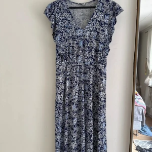 Blåvit långklänning från Indiska - Supersöt långklänning från Indiska i blått och vitt med blommigt mönster. Klänningen har v-ringning, korta ärmar och ett flowy fall. Perfekt för dig som gillar boho-vibes och vill ha något bekvämt men ändå snyggt.