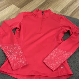 Rosa tränings-tröja SOC half zip - Sportig långärmad tränings-tröja från SOC i stark rosa med coola mönstrade detaljer på ärmarna. Tröjan har half zip framtill och är gjord i ett syntetmaterial som andas, perfekt för aktiva dagar. Snygg passform och riktigt bekväm att ha på sig.