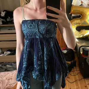 Blå mönstrad ärmlös linnetopp - Supersöt mörkblå linnetopp med turkost blommönster och smock upptill. Toppen har tunna axelband och en flowy, asymmetrisk nederdel som ger en bohemisk vibe. Perfekt för varma dagar och festivaler.