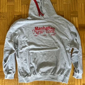 Grå hoodie Manhattan New York Carla Mara - Grå hoodie från Carla Mara med röd text 'Manhattan New York' och röda snören i huvan. Klassisk passform med muddar vid ärmslut och nederkant. Perfekt för dig som gillar streetstyle och vill ha en chill look.