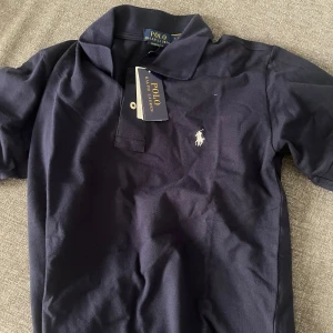 Marinblå pikétröja Polo Ralph Lauren - Snygg marinblå pikétröja från Polo Ralph Lauren i custom slim fit. Klassisk krage, kort ärm och ikoniska broderade loggan på bröstet. Tillverkad i bomull för en skön känsla och stilren look. Perfekt för dig som gillar preppy vibes.