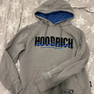 Grå hoodie från Hoodrich med blå detaljer - Snygg grå hoodie från Hoodrich med svart och blå logga på bröstet och ryggen. Fodrad huva med blå insida, känguruficka och cool streetwear-patch nertill. Långärmad och med snörning i huvan. Perfekt för dig som gillar streetstyle.