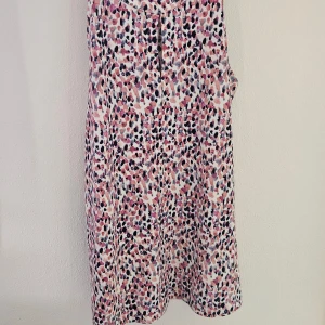 Ärmlös topp - Snygg ärmlös topp i rosa, svart och lila toner på vit botten. Klänningen har en liten ståkrage med guldfärgade knappar i nacken och en lätt A-linjeformad siluett. Perfekt för dig som gillar färg och mönster!