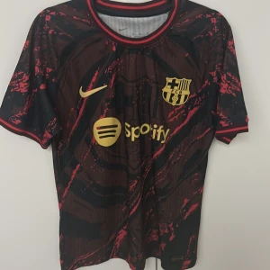 FC Barcelona Nike x Spotify matchtröja - Snygg FC Barcelona matchtröja från Nike med Spotify-logga på bröstet. Tröjan är svart med röda detaljer och har ett unikt abstrakt mönster. Korta ärmar och rund hals med röd och svart ribbad kant. Materialet är lätt och andas, perfekt för fotboll eller streetwear.
