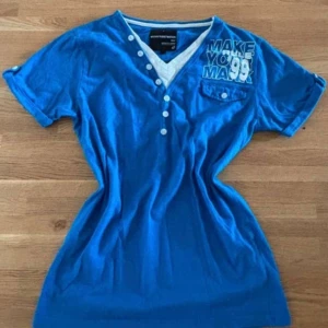 Blå t-shirt Outfitters Nation - Blå t-shirt från Outfitters Nation med vit texttryck och coolt #99-motiv. T-shirten har v-ringning med knappar, korta ärmar och en liten bröstficka med knapp. Materialet är mjuk bomull och passformen är normal. Perfekt för dig som gillar sportig och avslappnad stil. Väldigt fin💕Har aldrig används av mig och finns inget stort tecken på användning av tidigare ägare💗🤗