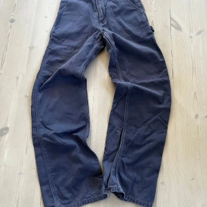Blå cargopants från Carhartt - Säljer ett par mörkblå cargopants från Carhartt med bred passform och flera praktiska fickor. Byxorna är tillverkade i slitstark bomull och har klassisk Carhartt-logga på bakfickan. Perfekta för dig som gillar streetwear och funktionella detaljer.