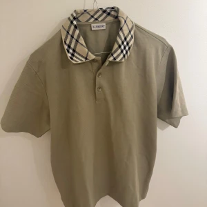 Grön Burberry pikétröja med rutig krage - Snygg mjuk-grön pikétröja från Burberry med klassisk rutig krage i beige, svart och vit. Tröjan har korta ärmar, tre knappar framtill och är gjord i bomull. Perfekt för dig som vill ha en stilren och tidlös look med en touch av lyx.