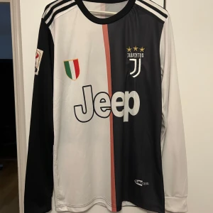 Juventus långärmad Ronaldo tröja - Juventus långärmad matchtröja med Ronaldo 7 på ryggen. Tröjan är svart och vit med en rosa rand i mitten, Juventus-emblem och Jeep-logga på bröstet samt Serie A-märke på ärmen. Tillverkad i lätt och ventilerande Climalite-material.