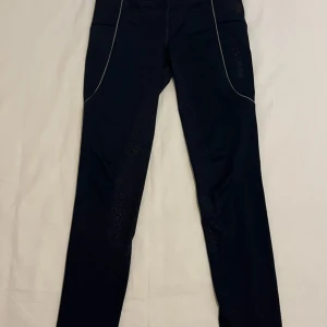 Mörkblå ridleggings från Kingston - Säljer ett par mörkblå ridleggings från Kingston med silikongrepp på insidan av benen och reflekterande detaljer längs sidorna. Byxorna har hög midja och är tillverkade i stretchigt syntetmaterial som ger en tight och bekväm passform. Perfekta för ridning.