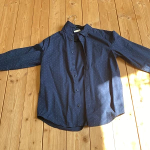 Mörkblå prickig skjorta Kappahl 152 - Snygg mörkblå skjorta från Kappahl i storlek 152. Skjortan har små vita prickar, klassisk krage och knäppning framtill. Tillverkad i mjuk bomull och har långa ärmar. Perfekt för dig som vill ha en stilren look med lite extra detaljer.