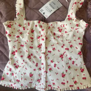 Blommig topp från ZARA - Supersöt korsettopp från ZARA i vit med små röda blommor, knappar framtill och volangdetaljer på axelbanden. Aldrig använd, med prislapp kvar! Originalpris=329kr❤️🌸