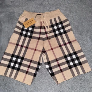 Burberry shorts  - Snygga shorts från Burberry i klassiskt beige rutigt mönster med svarta, vita och röda detaljer. De har elastisk midja med snörning och är tillverkade i ett mjukt stickat material. Perfekta för dig som vill ha en lyxig och trendig vibe.