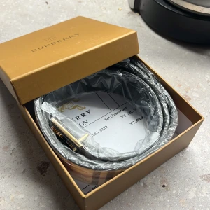 Burberry rutigt bälte med B-spänne - Snyggt Burberry-bälte med klassiskt rutig design i beige, svart, rött och vitt. Bältet har ett stort metallspänne med Burberrys ikoniska B-logga i guld och svart. Perfekt accessoar för att lyfta din outfit med en lyxig touch.