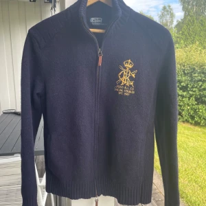Mörkblå Zip-tröja från Polo Ralph Lauren - Mörkblå kofta i ull från Polo Ralph Lauren med gul broderad logga och text på bröstet. Tröjan har hel dragkedja, ribbade muddar och hög krage. Inga fläckar eller skador. Mindre i storlek. säljer pga för liten storlek. 