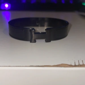 Svart armband med H-spänne - Stilrent svart armband med ett coolt H-format spänne framtill. Armbandet har en enkel och modern design som passar perfekt till streetwear eller en mer clean look. Tillverkat i metall med en blank finish som ger en snygg touch till din outfit.