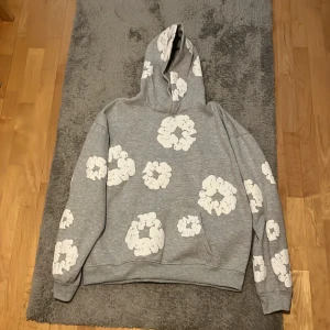 Grå hoodie med vita blommor Denim Tears - Säljer en grå hoodie från Denim Tears i storlek L. Tröjan har stora vita blommönster över hela plagget, huva och långa ärmar. Mjuk insida och relaxed fit, perfekt för dig som gillar streetwear med unik design.