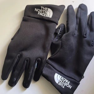 Svarta handskar The North Face - Svarta, smidiga handskar från The North Face med vit logga på ovansidan. Handskarna är tunna men ändå varma, perfekta för kyliga dagar. Snygg minimalistisk design med åtsittande passform och mjukt syntetmaterial.