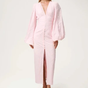 Ljusrosa långklänning med puffärm från adoore - Superfin ljusrosa långklänning med djup v-ringning, puffiga långärmar och knappar framtill. Använd endast en gång 